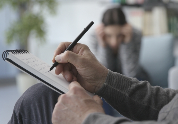 Psychologist taks anamnesis of a patient preparing the treatment for depression. The picture is showing a person sitting in the chair doing notes in a notbook. There is a blurred silhouette of a patient in the background.