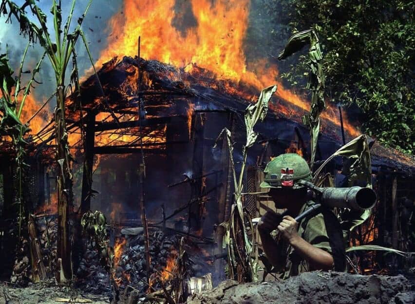 PTSD combat trauma in Vietnam war. The picture reflects the Vietnam war trauma affecting soldiers and civilians. In the front of the picture an American soldier in combat dress is carrying a mortar. In the background burning Vietnamese wooden house in the middle of a jungle. The Vietnam war initiated intense research on PTSD