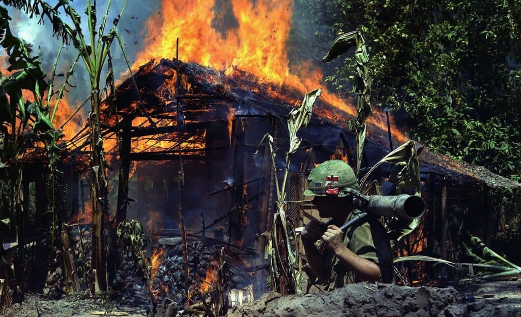 The picture reflects the Vietnam war trauma affecting soldiers and civilians. In the front of the picture an American soldier in combat dress is carrying a mortar. In the background burning Vietnamese wooden house in the middle of a jungle. The Vietnam war initiated intense research on PTSD