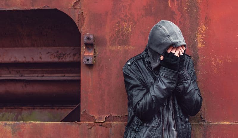 A young man in a black leather jacket wearing a gray hoody.  He has his face covered with his hands that are clad in gloves with cut off fingers.  He is standing in front of a reddish, rusty industrial machine.  This image represents the shame and guilt addicts suffer when they are still active in their dependency.  Many addicts become socially isolated and are not able to hold jobs.  