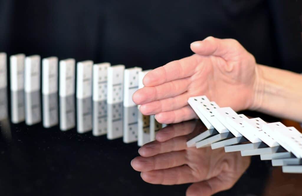 Domino stones that are lined up with a hand separating two parts of the line-up on a black background.  The one side is at the back of the hand shows upright dominos.  The other side has fallen against the palm of the hand.  The hand represents stopping the domino effect of relapse triggers adding up to an inevitable relapse.  Good relapse prevention stops the relapse progression.    