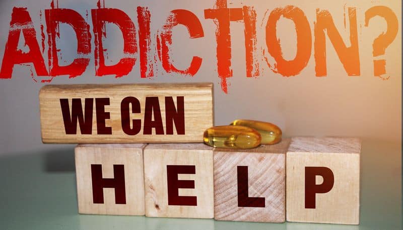 The Word addiction in capitalized red letters with a question mark written above the words, we can, that written on a piece of wood.  Below that the word Help spelled out on wooden blocks.  There are two gold colored capsules of  a golden color.