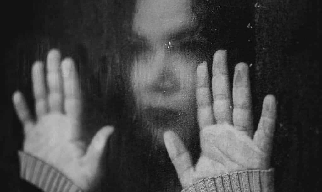A black-and-white picture of a woman who is pushing her hands against a wet window pane.  This shows the sadness and isolation of dependency and addiction.  