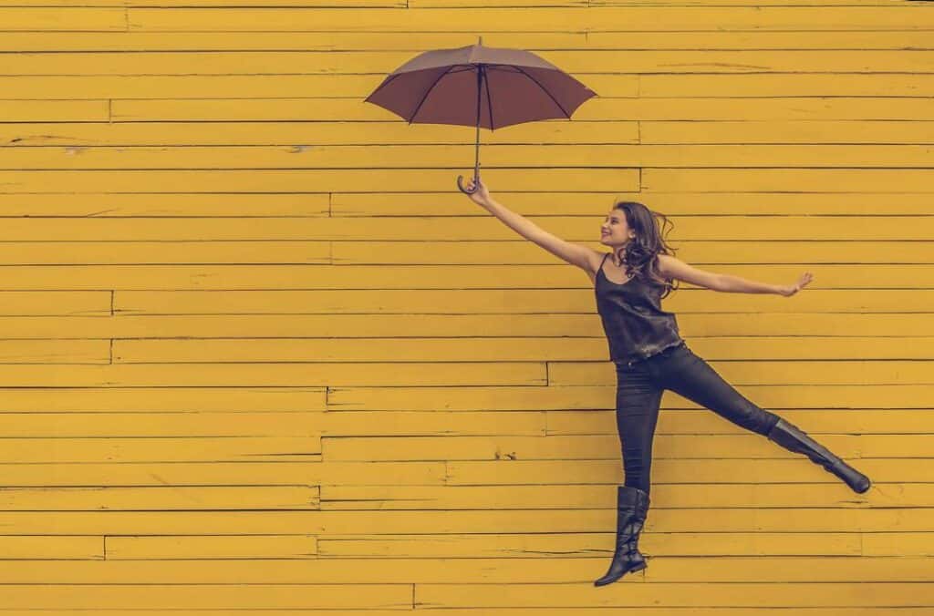 A woman holding up an umbrella while dancing, on yellow background, expressing joy. Quality of life is reinstated when people overcome the stigma of psychiatry and mental healthcare and get help for their mental health disorders.  