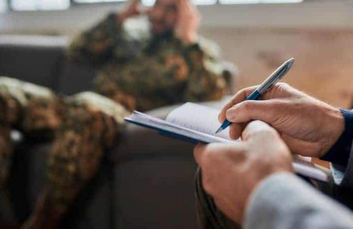 PTSD treatment in Dubai. Psychotherapy (counselling) for PTSD in Dubai. The picture shows the psychotherapy treatment for PTSD. The soldier in a field uniform is half lying on the sofa. In front of him we see the arms of the psychotherapist holding a pen and making notes

