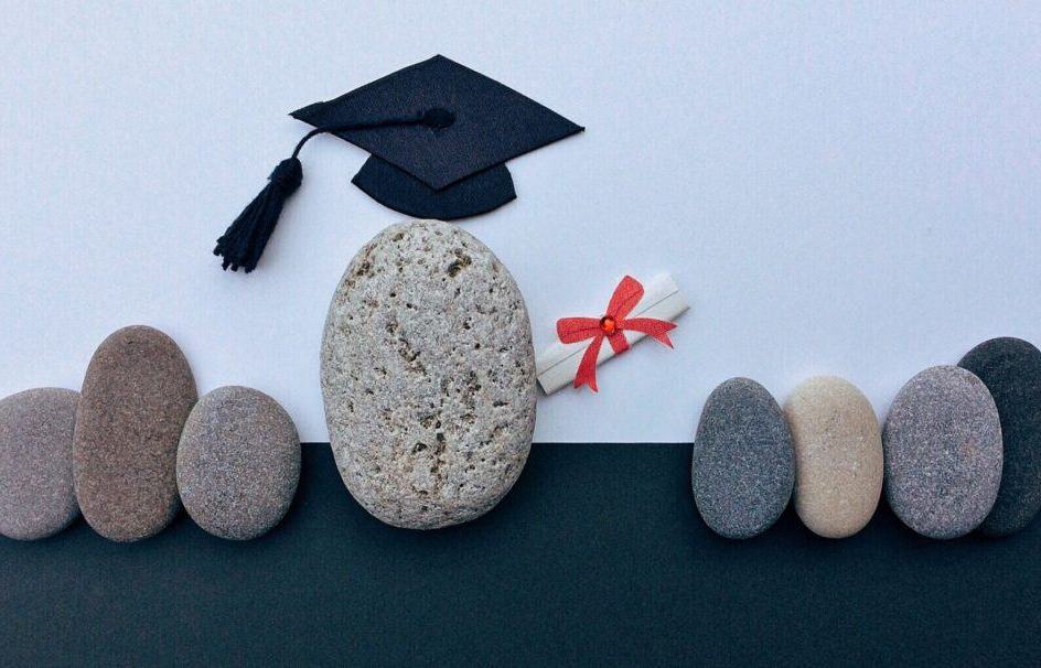 Best psychologist Dubai. There are 8 natural stones in a row on a black surface with a light blue background. They are mostly the same small size with the exception of the 4th one from the left.  That stone is larger and has a graduate cap on it, and one arm visible on the right side made out of a paper scroll with a red ribbon on it.  This stone represents a university graduate.  The best psychologist is Dubai is one that has university credentials.  