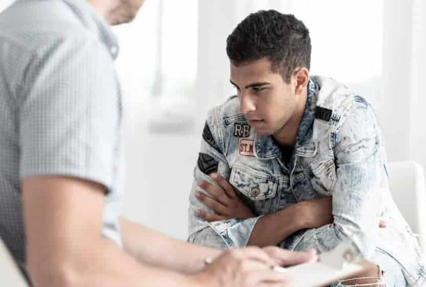 Dependency Counselling. Young man sitting in front of a psychotherapist. The man bends forward, his arms are crossed. We can see only the shoulder and the right arm of the psychotherapist. Counselling for addiction