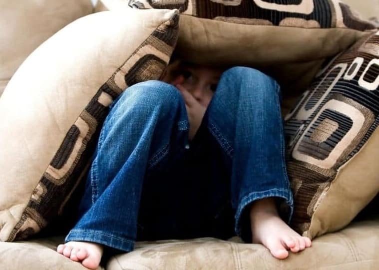 Major Depressive Disorder (MDD). Social withdrawal. A teenager sitting on the light brown sofa covering the body with pillows. In the depth we see only a part of the depressed face. The picture shows how depression affects the social functioning with lack of self-esteem and social withdrawal